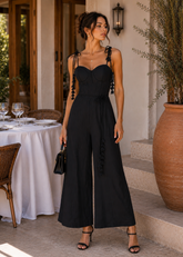 Woman in a black jumpsuit standing in an elegant outdoor setting with a table and chairs.