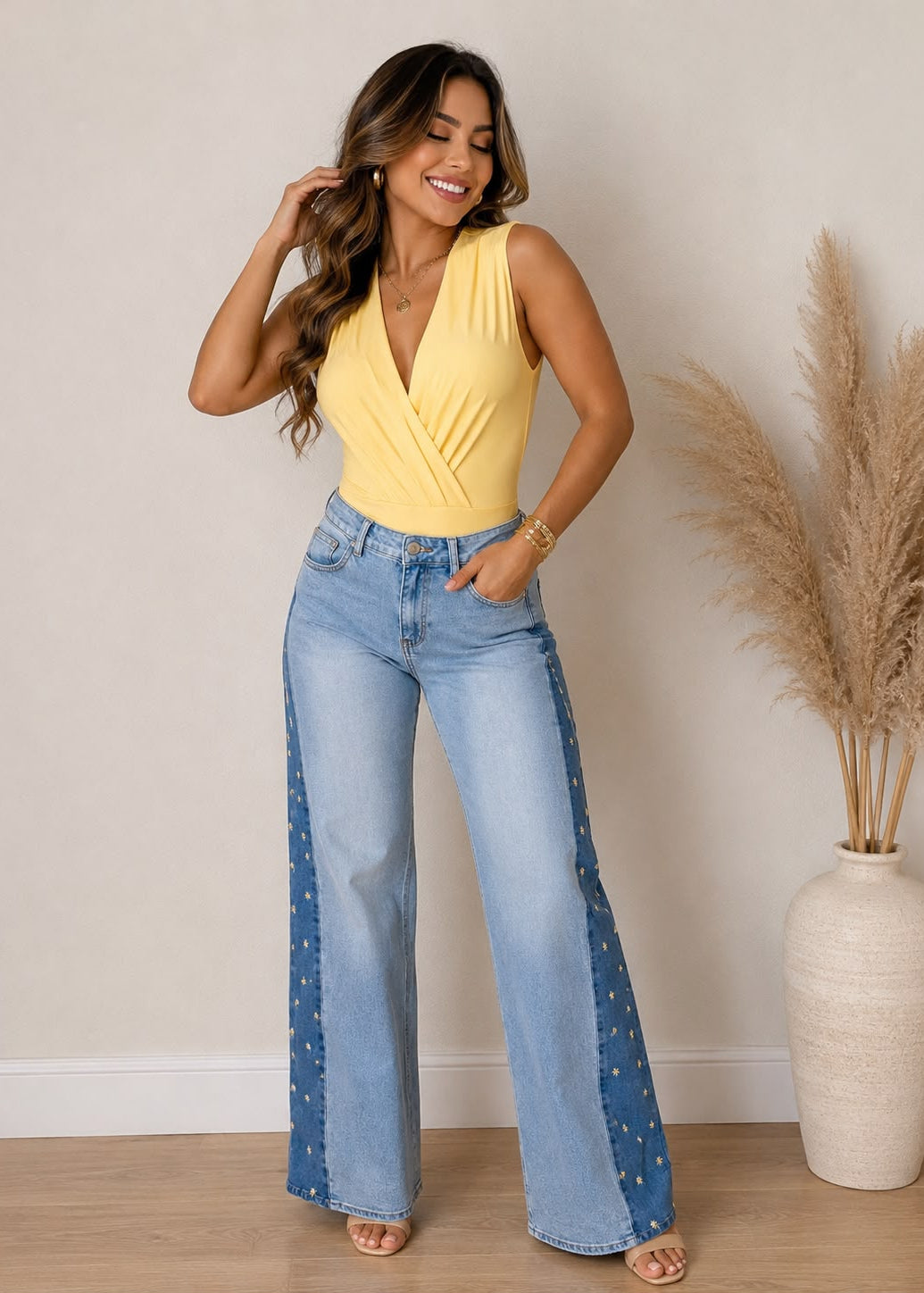 Woman wearing a yellow top and blue jeans standing against a neutral wall with a vase of dried plants.