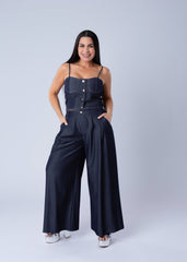 A woman modeling a dark navy blue denim jumpsuit with wide-leg pants, a buttoned bodice, and thin shoulder straps, standing against a light gray studio background.